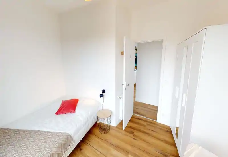 Dormitorio individual compacto y luminoso con armario blanco y suelo de madera. La cama está bien arreglada con un cojín rojo y una mesita, ideal para una estancia privada y acogedora.