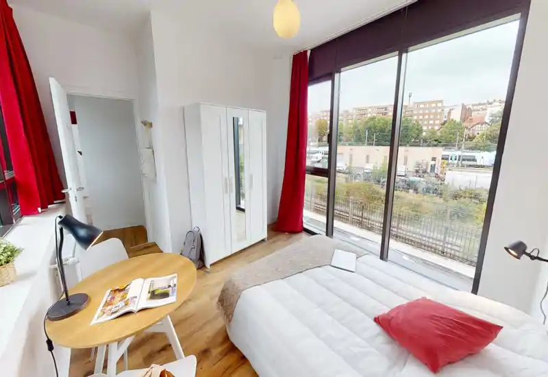 Bright, modern bedroom with large floor-to-ceiling windows, a comfortable double bed with a red cushion, white wardrobe and a small workspace beside the window.