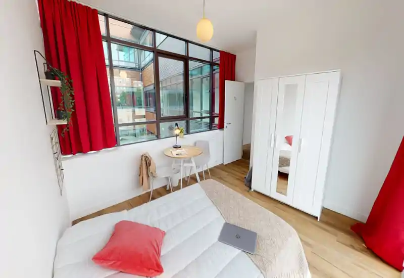 Cozy, bright single bedroom with a neatly made bed, red curtains and a large industrial-style window that fills the room with natural light. A small round table and chairs by the window and a white wardrobe complete the practical layout.
