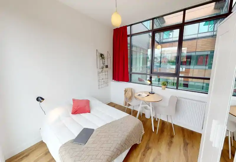 Bright modern single bedroom with a neatly made double bed, wooden floor and large floor-to-ceiling window providing abundant natural light. A small round table with chairs and minimal decorative accents create a cozy, functional space ideal for a short-term stay.