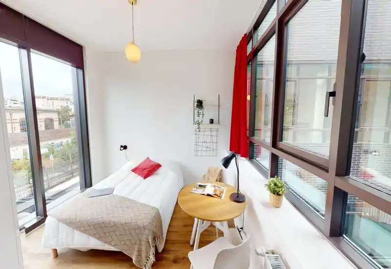 Bright modern bedroom with large floor-to-ceiling windows, a neatly made double bed with warm textiles and a small round table by the window — ideal for work or reading with lots of natural light.