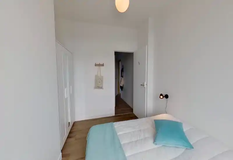 Dormitorio sencillo y luminoso con cama blanca, ropa de cama clara y una manta y cojín turquesa. Tiene armario, suelo de madera e iluminación moderna que aportan sensación de orden y amplitud.