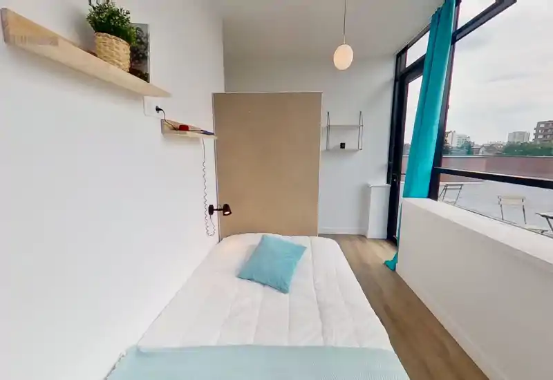 Camera da letto compatta e luminosa con letto ordinato, accenti turchesi e ampia finestra che offre molta luce naturale e vista sul balcone.