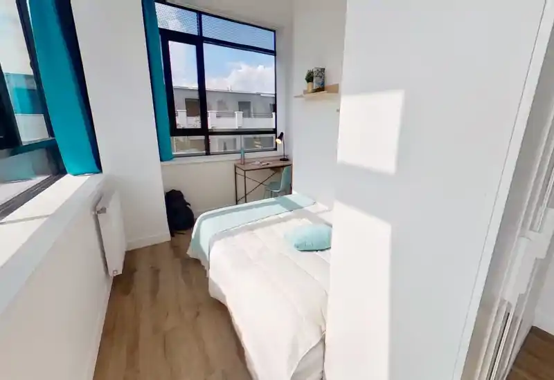 Camera da letto singola luminosa e moderna con letto ordinato, dettagli turchesi e una scrivania compatta vicino a una grande finestra che illumina la stanza.