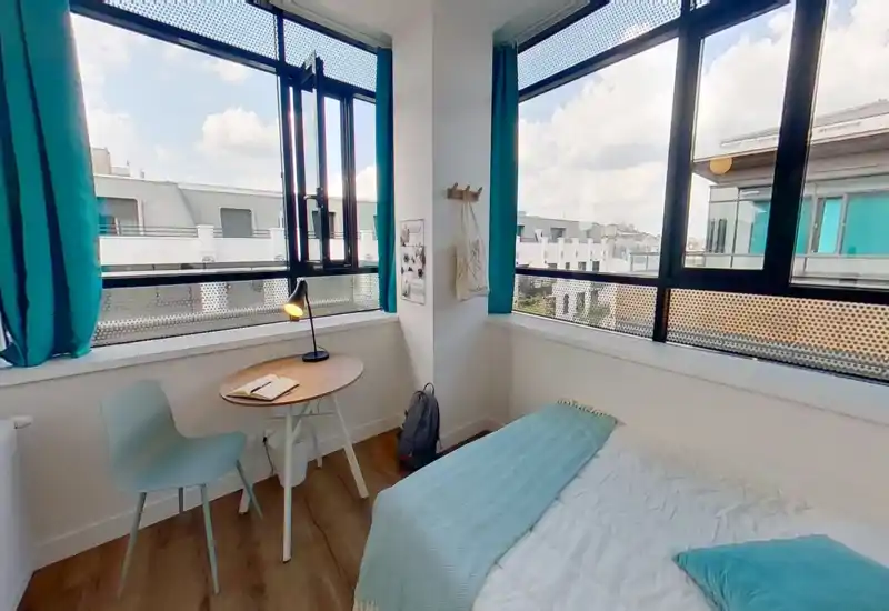 Dormitorio moderno y luminoso en esquina con grandes ventanas, cama cómoda con manta y cojín, y pequeño escritorio con lámpara — perfecto para descansar o estudiar con mucha luz natural.