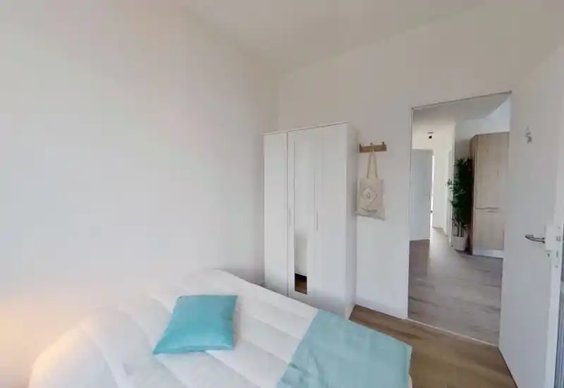 Minimal, bright bedroom with a white double bed dressed in light textiles and a turquoise accent pillow and throw; a white wardrobe and open doorway lead to a hallway with plant accents.