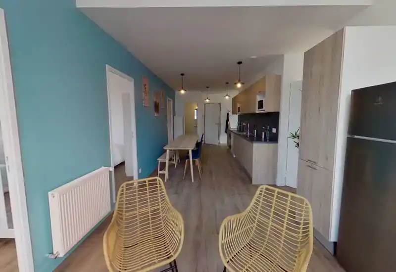 Bright, modern open-plan living area with a long dining table, kitchen units along one wall and two rattan lounge chairs in the foreground — ideal for social dining and relaxed evenings.