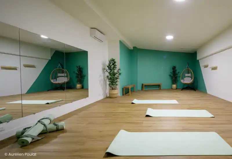 Bright, modern living area with wooden floors, decorative plant and floor mats; suitable for relaxing or light exercise. The image shows a long open space with clean lines and soft lighting.