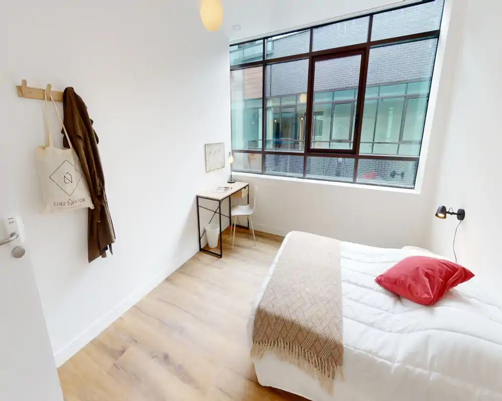 Dormitorio luminoso y minimalista con una cama cómoda, manta cálida y cojín rojo. Gran ventana que aporta mucha luz natural y un escritorio compacto para trabajar.