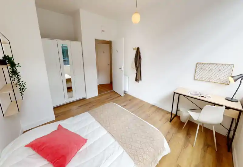 Dormitorio moderno y luminoso con cama bien preparada, suelo de madera y un pequeño escritorio junto a la ventana, ideal para una persona o estudiante. Paredes blancas y decoración minimalista que crean un ambiente fresco y aireado.