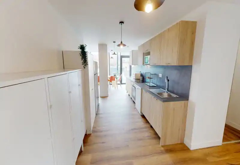 Cocina moderna en galería, luminosa, con muebles efecto madera, electrodomésticos integrados y fregadero a la derecha; amplias ventanas al fondo proporcionan luz natural.