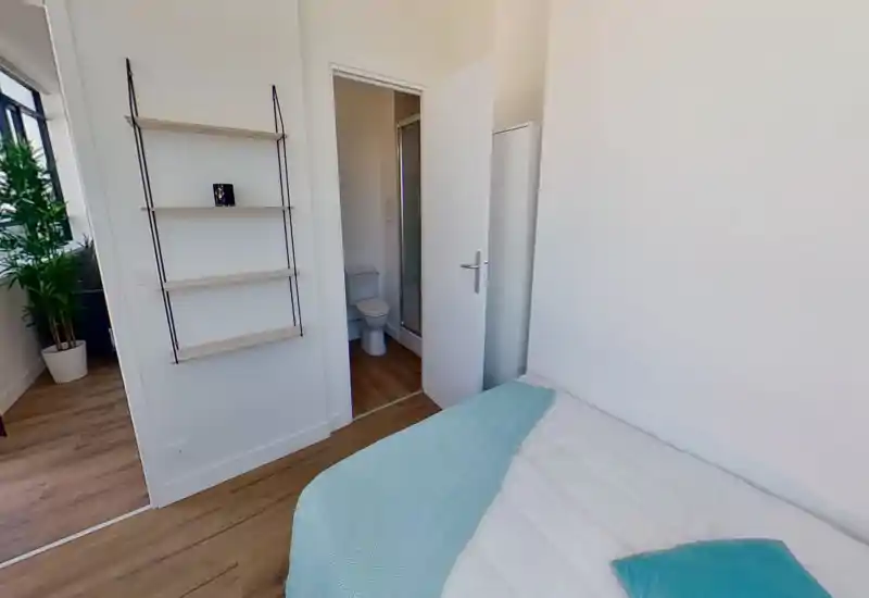 Dormitorio luminoso y minimalista con cama blanca y colcha turquesa; colchón y almohada claramente visibles y baño en suite accesible por la puerta abierta.