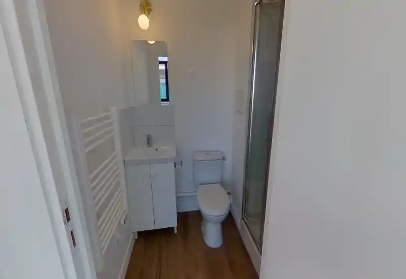 Baño compacto y moderno con lavabo y mueble, inodoro y mampara de ducha de vidrio. Paredes blancas y suelo efecto madera para un aspecto limpio y contemporáneo.