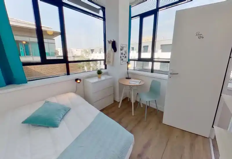 Dormitorio individual luminoso y moderno con cama cómoda, luz de mesilla y una pequeña mesa junto a grandes ventanas que proporcionan mucha luz natural.