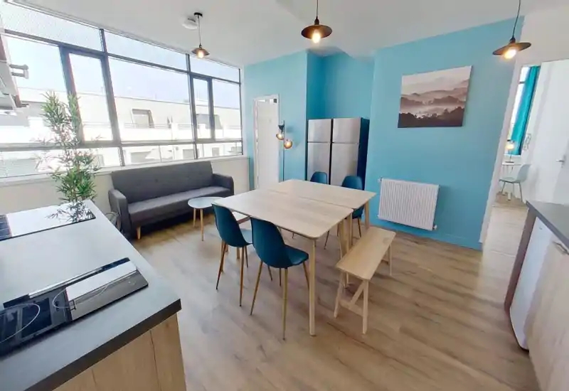 Amplia zona de estar/comedor luminosa y moderna con grandes ventanales, sofá gris, mesa de madera central con sillas azules y lámparas colgantes contemporáneas.