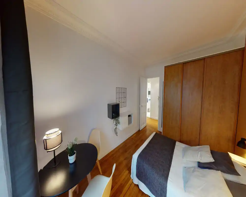 Chambre lumineuse et bien décorée avec lit double, parquet chaleureux et grands placards muraux en bois. Petite table avec chaises et lampes de chevet apportent confort et fonctionnalité.