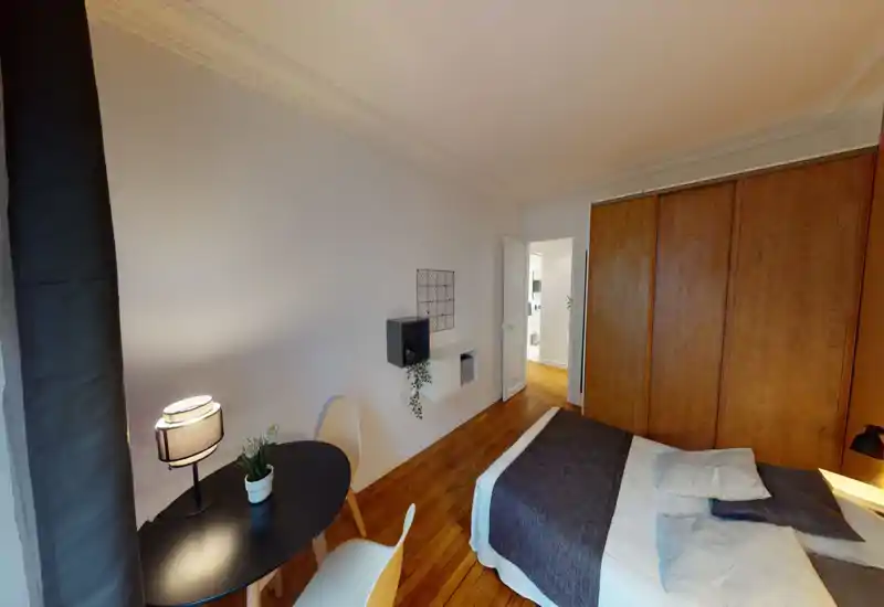 Chambre lumineuse et bien décorée avec lit double, parquet chaleureux et grands placards muraux en bois. Petite table avec chaises et lampes de chevet apportent confort et fonctionnalité.