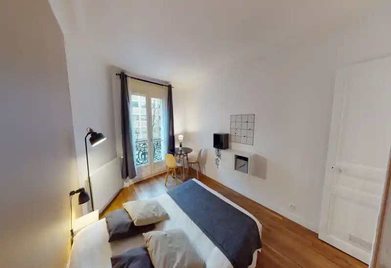 Chambre lumineuse et soignée avec un lit double au premier plan, éclairage d'appoint chaleureux et un petit coin table près de la fenêtre — idéale pour un séjour citadin confortable.