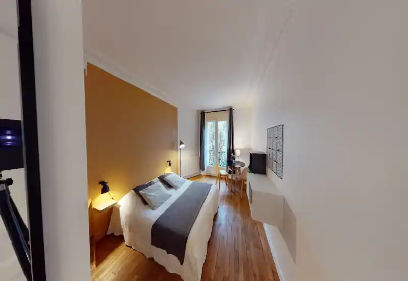 Chambre lumineuse et élégante avec un grand lit double contre un mur ocre, lampes de chevet et un petit coin table/préparation près de la fenêtre. Parquet et moulures au plafond apportent du caractère.