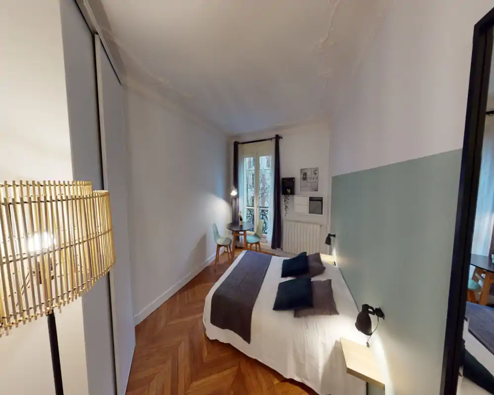 Chambre lumineuse et élégante avec lit double, textiles doux et parquet en chevrons. Une petite table avec chaises près de la fenêtre et des lampes chauffantes créent une atmosphère chaleureuse.