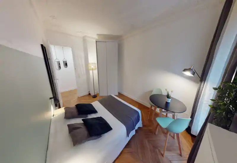 Chambre lumineuse et élégante avec un lit double, coussins décoratifs et une petite table près de la fenêtre. Parquet en chevrons et éclairage doux pour une atmosphère chaleureuse et contemporaine, idéale pour la location courte durée.