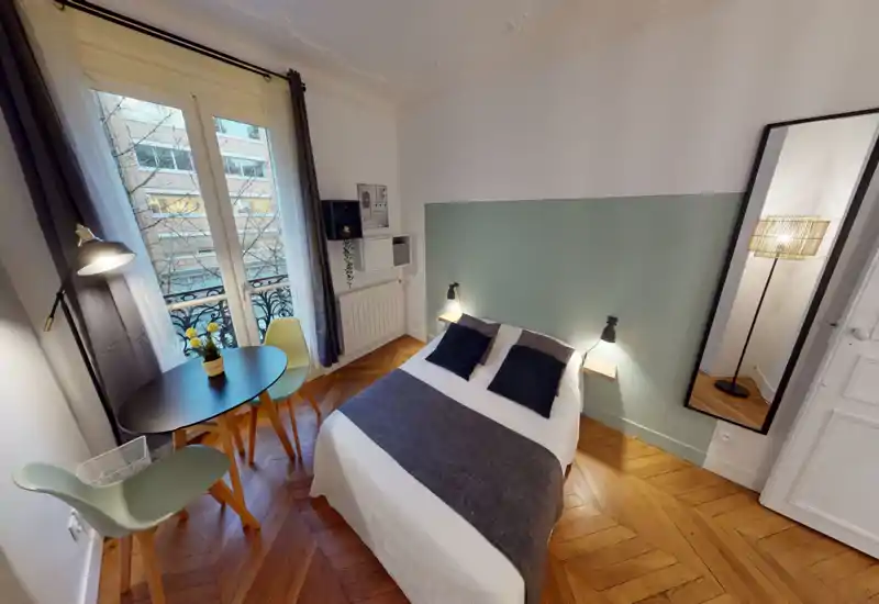 Chambre lumineuse et élégante avec un lit double devant un mur vert pâle, parquet en chevrons et petite table près de la fenêtre — idéale pour un séjour confortable en ville.