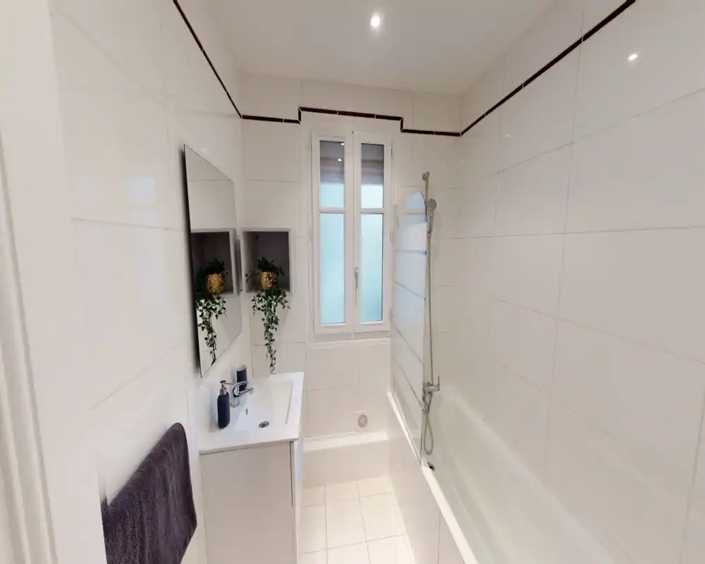 Salle de bains moderne et lumineuse avec baignoire et pare-douche, miroir mural et lavabo meuble. Carrelage blanc et lumière naturelle par la fenêtre pour une atmosphère fraîche et aérée.