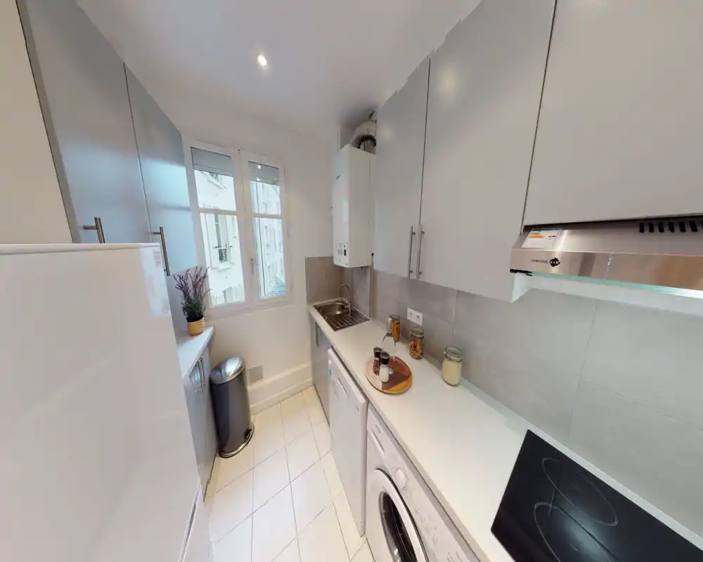 Cuisine moderne et compacte, lumineuse avec des meubles blancs, plan de travail et plaque de cuisson intégrée. Agencement propre et fonctionnel, adapté aux appartements urbains.