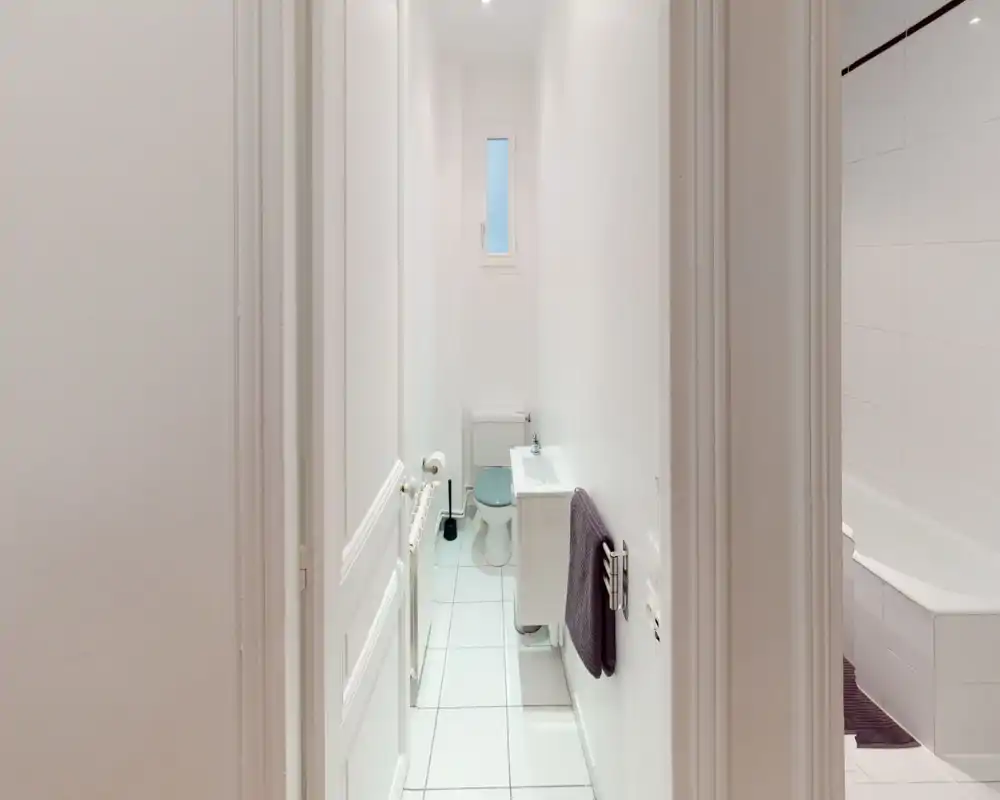 Salle de bain étroite et moderne avec carrelage blanc, WC, petit lavabo et baignoire visible par l’ouverture ; lumineuse, propre et fonctionnelle.