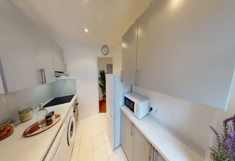 Cuisine compacte et moderne avec placards clairs, électroménagers intégrés et plan de travail lumineux — bien éclairée et soigneusement décorée avec bocaux et micro-ondes.