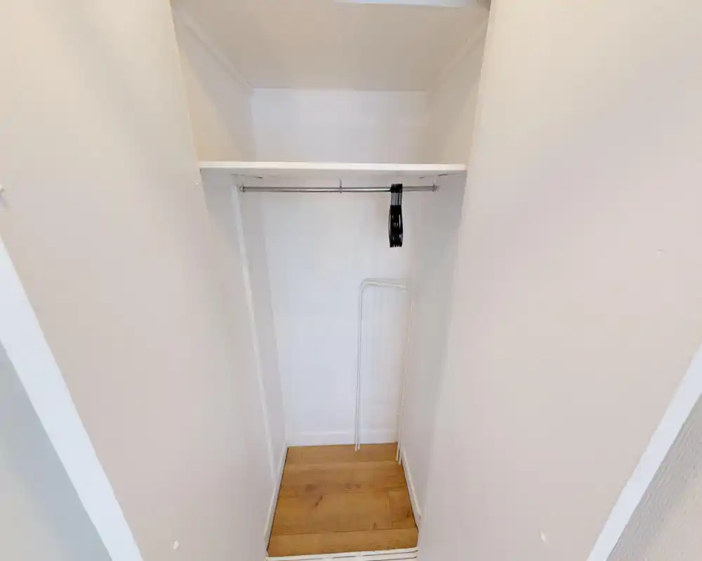 Small built-in closet with a hanging rail and a shelf, clean white walls and wood-effect floor — ideal for coats and storage.