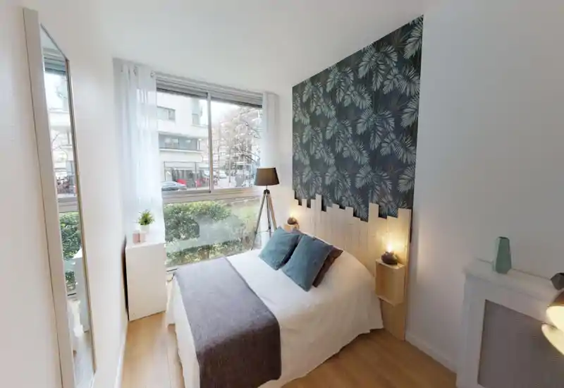 Bright, well-styled bedroom with a double bed facing a large window, decorative leaf-patterned feature wall and warm bedside lighting — ideal for a cozy city stay.
