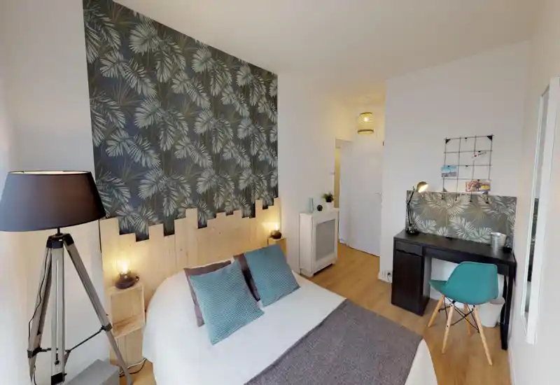 Bright, stylish bedroom with a double bed, patterned accent wall and cozy textiles. A bedside lamp and floor lamp create warm ambient light, while a desk area offers a practical workspace.