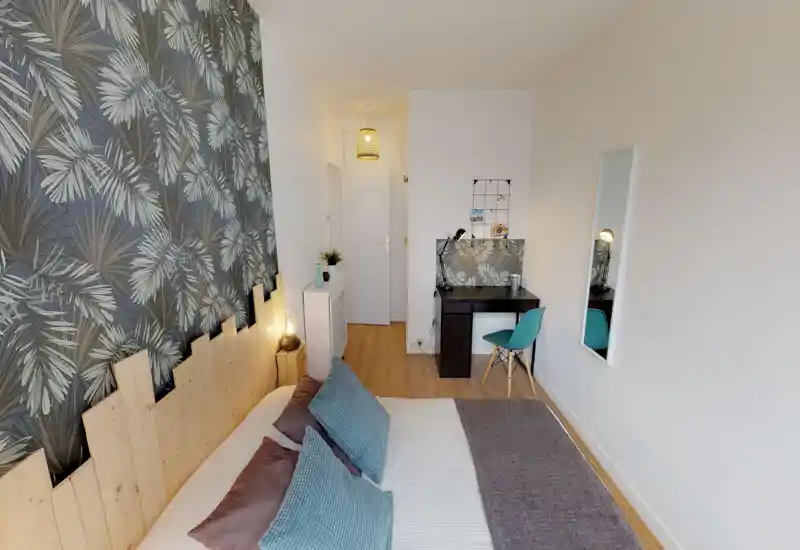 Bright, stylish bedroom with a large bed in the foreground, decorative tropical wallpaper and a compact desk area — cozy and well lit for relaxing or working.