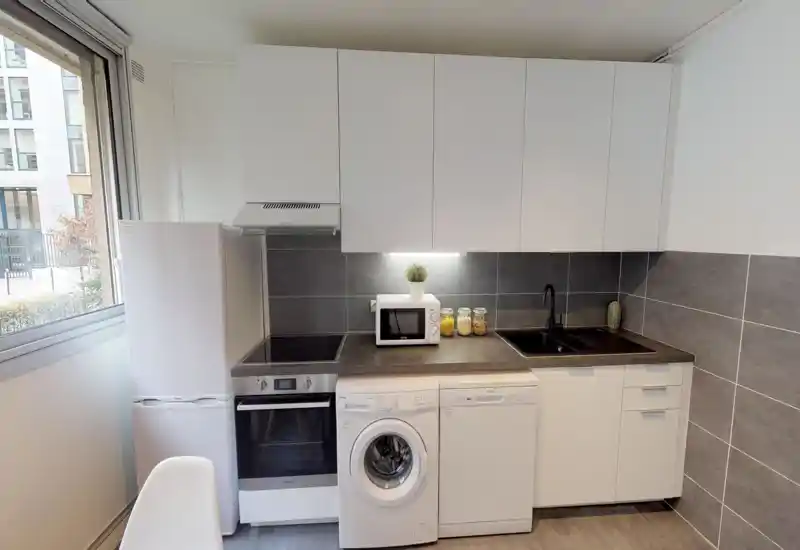 Modern compact kitchen with integrated appliances: fridge, oven, washing machine and dishwasher under a dark countertop, white cabinets and a sink with black faucet. Bright window light and neat finishes create a clean, functional space ideal for city apartments.