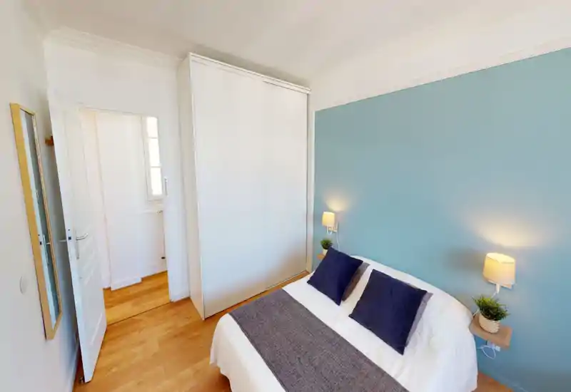 Bright, tidy bedroom with a double bed dressed in white linens, navy cushions and a gray runner; built-in white wardrobe and soft blue accent wall create a calm, modern feel.