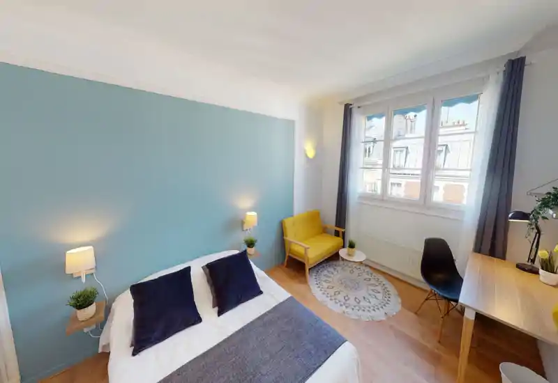 Bright, modern bedroom with a large bed, soft textiles and a cheerful yellow loveseat by the window. The room features a calming blue accent wall, wooden flooring and a desk area ideal for work.
