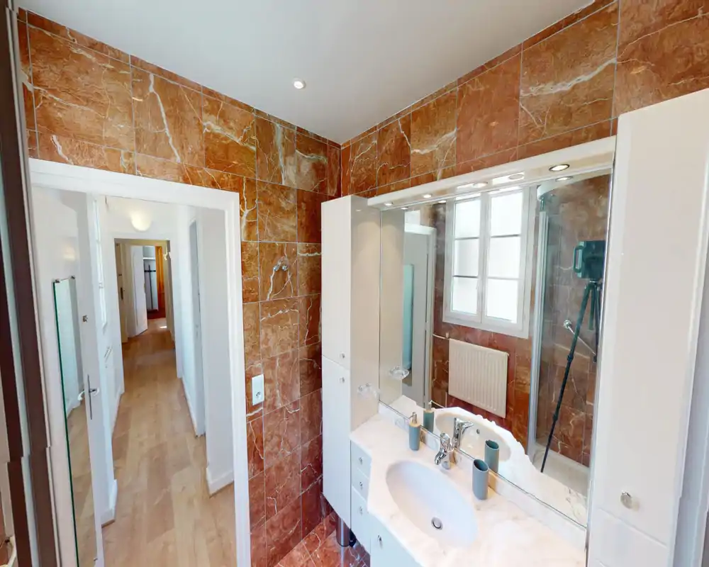 Bright, tiled bathroom with marble-effect walls, a white vanity with sink and mirror, and a glass shower enclosure. The space is well lit and finished with modern fixtures.