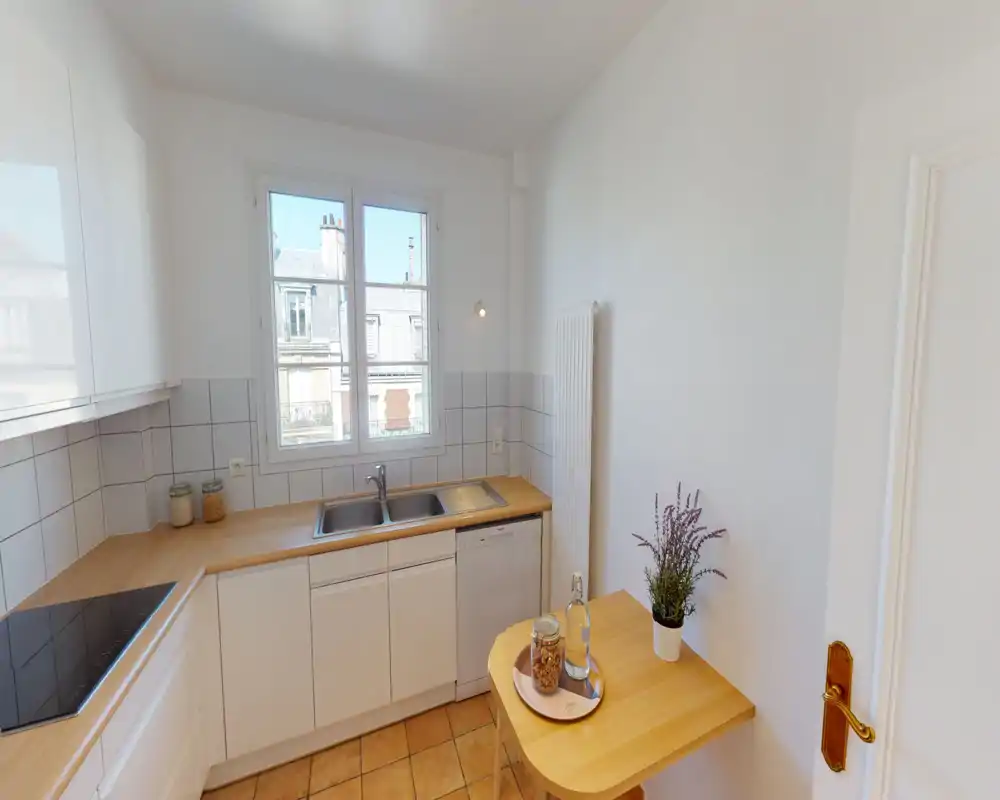 Compact, bright kitchen with white cabinets, wooden countertops, sink under a window and a small breakfast table with a plant — tidy and well lit by natural light.
