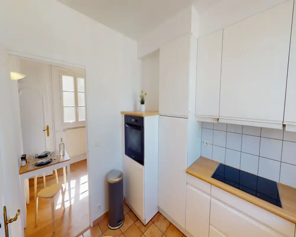 Bright, compact kitchen with white cabinets, wooden countertops, built-in oven and glass-ceramic hob. The space is neat and well lit, with a small plant adding a homely touch.