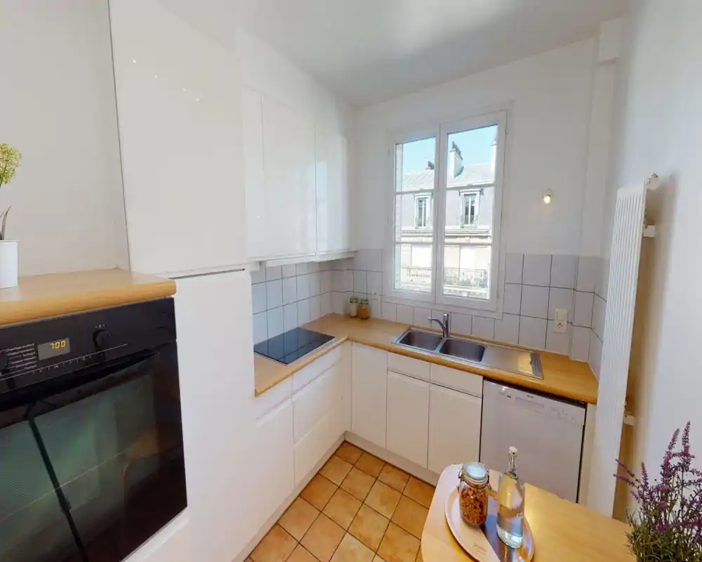 Compact, bright kitchen with white cabinetry and warm wooden countertops. Includes an induction cooktop, built-in oven, double sink and dishwasher; a window provides natural light and a pleasant view.