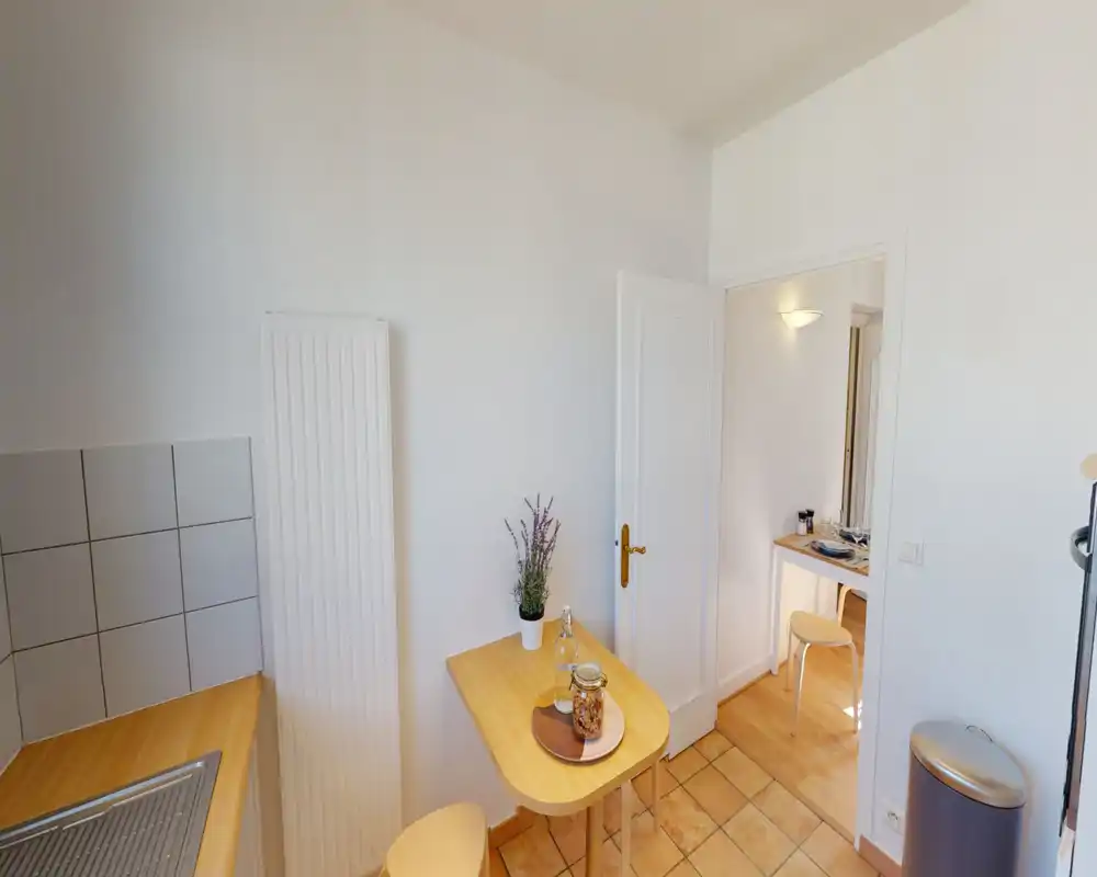 Compact, well-lit kitchen corner with a wooden breakfast table, stools and a sink area. Neutral white walls and warm wooden surfaces create a tidy, inviting space ideal for casual meals.
