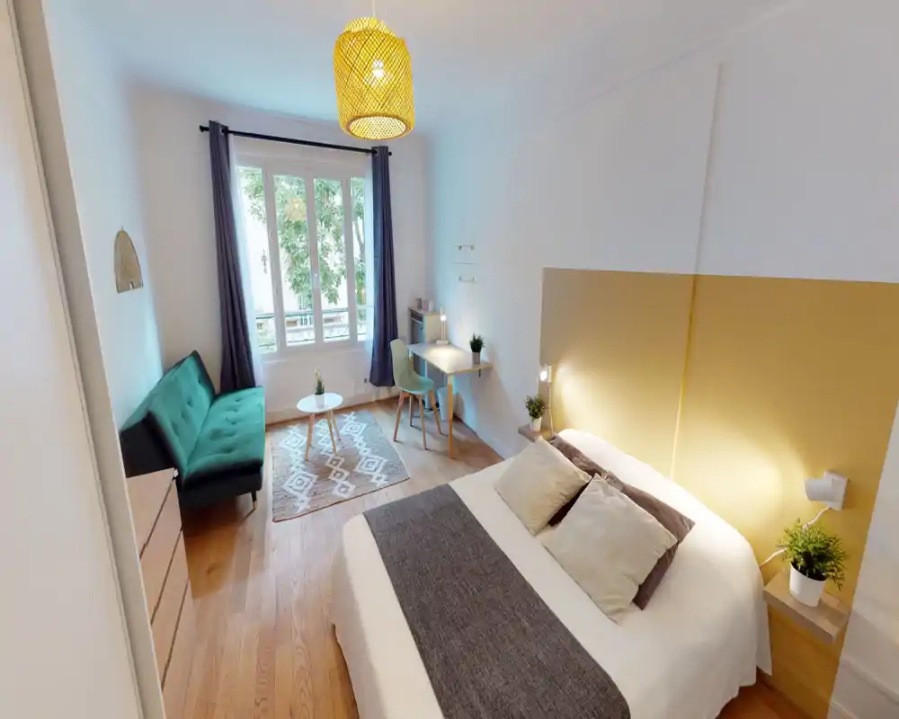 Bright, well-styled bedroom with a comfortable double bed, bedside lamps, a small desk and a green sofa by the window — ideal for a calm, cozy stay.