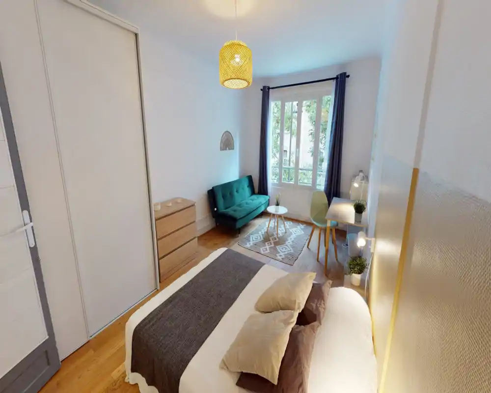 Bright, well-styled bedroom with a comfortable double bed in the foreground, a green sofa and desk by the window, wooden floors and warm lighting — ideal for a cozy city stay.