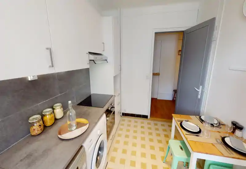 Compact, well-equipped kitchen with white cabinets, a ceramic cooktop, countertop appliances and a small dining nook set for two — bright and tidy space ideal for short stays.