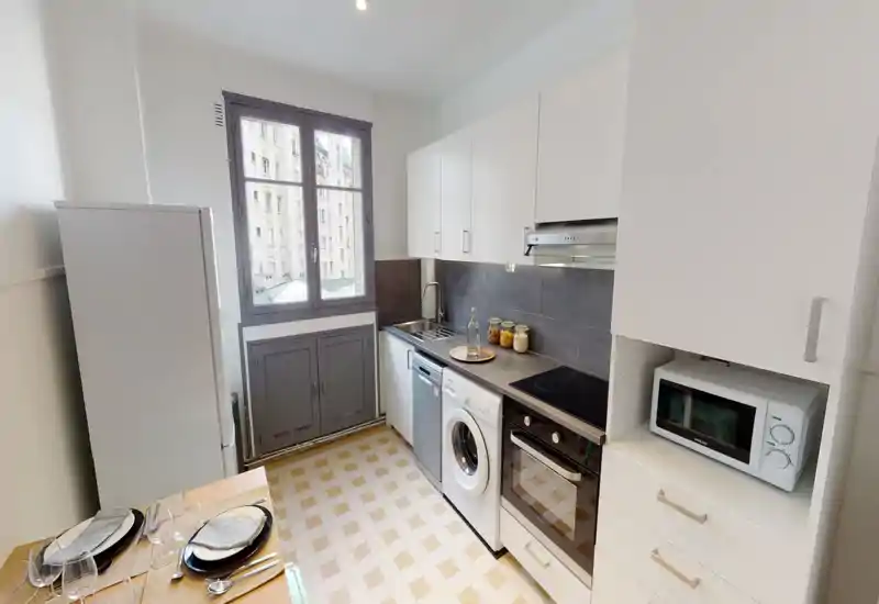 Cuisine compacte et lumineuse avec des placards blancs modernes, plaque de cuisson électrique, four, micro-ondes et lave-linge intégré. La grande fenêtre apporte de la lumière naturelle sur un plan de travail soigné avec des bocaux décoratifs et un coin repas.