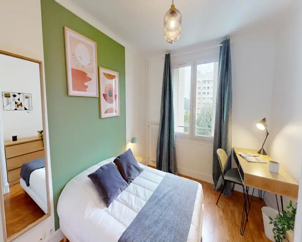 Camera da letto luminosa e curata con parete verde, letto matrimoniale confortevole, lampada da comodino e una scrivania compatta vicino alla finestra — ambiente tranquillo e accogliente.