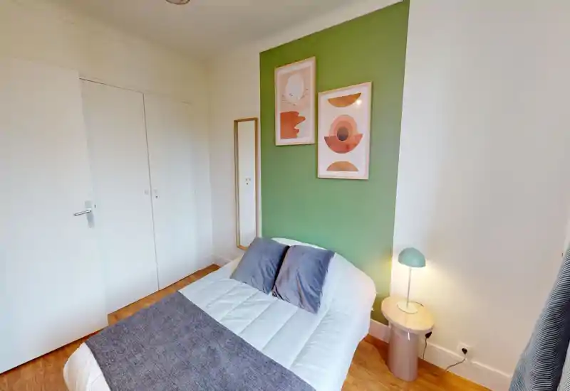 Camera da letto luminosa e moderna con letto matrimoniale davanti a una parete verde, cuscini coordinati e quadri; pavimento in legno e lampada da comodino completano l'ambiente accogliente.