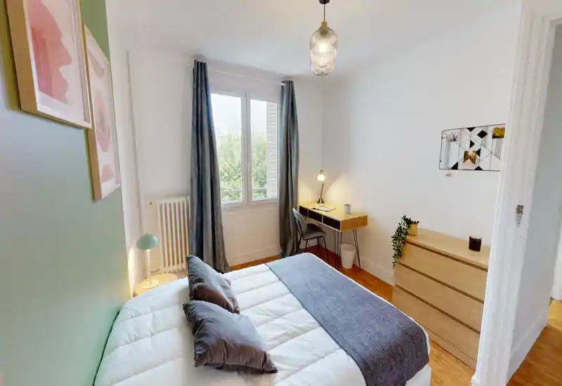 Camera da letto moderna e luminosa con un comodo letto matrimoniale in primo piano, una scrivania vicino alla finestra e un comò in legno. La luce naturale, i tessili scelti con gusto e l’arredamento essenziale rendono l’ambiente accogliente e funzionale.