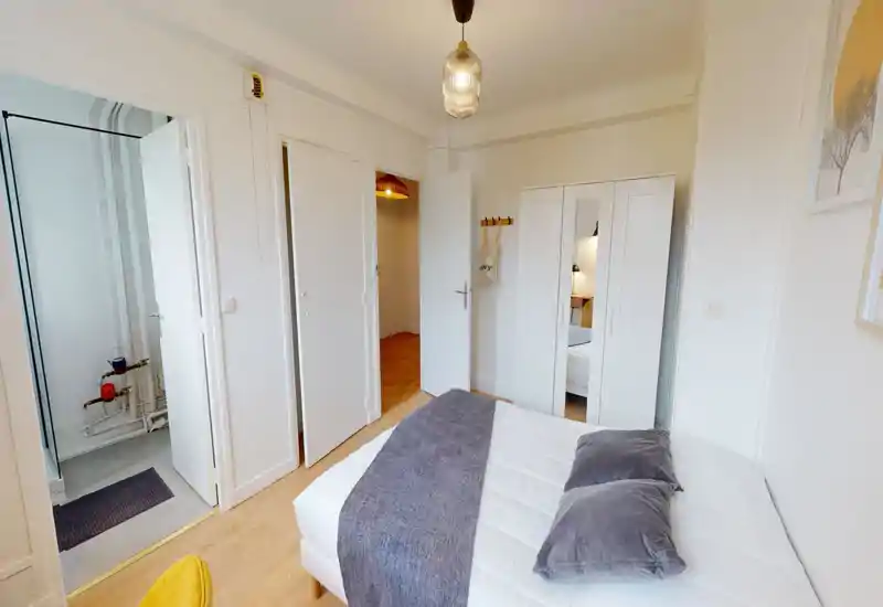 Chambre lumineuse et soignée avec un grand lit bien fait, coussins gris et jeté assorti ; armoire blanche avec miroir et sol en bois clair pour une atmosphère propre et accueillante.